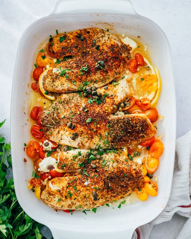 a white casserole dish with chicken, tomatoes and lemons on the side