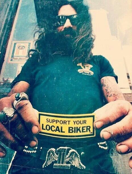 a man with long hair and beard wearing a black shirt holding a yellow label that says support your local biker