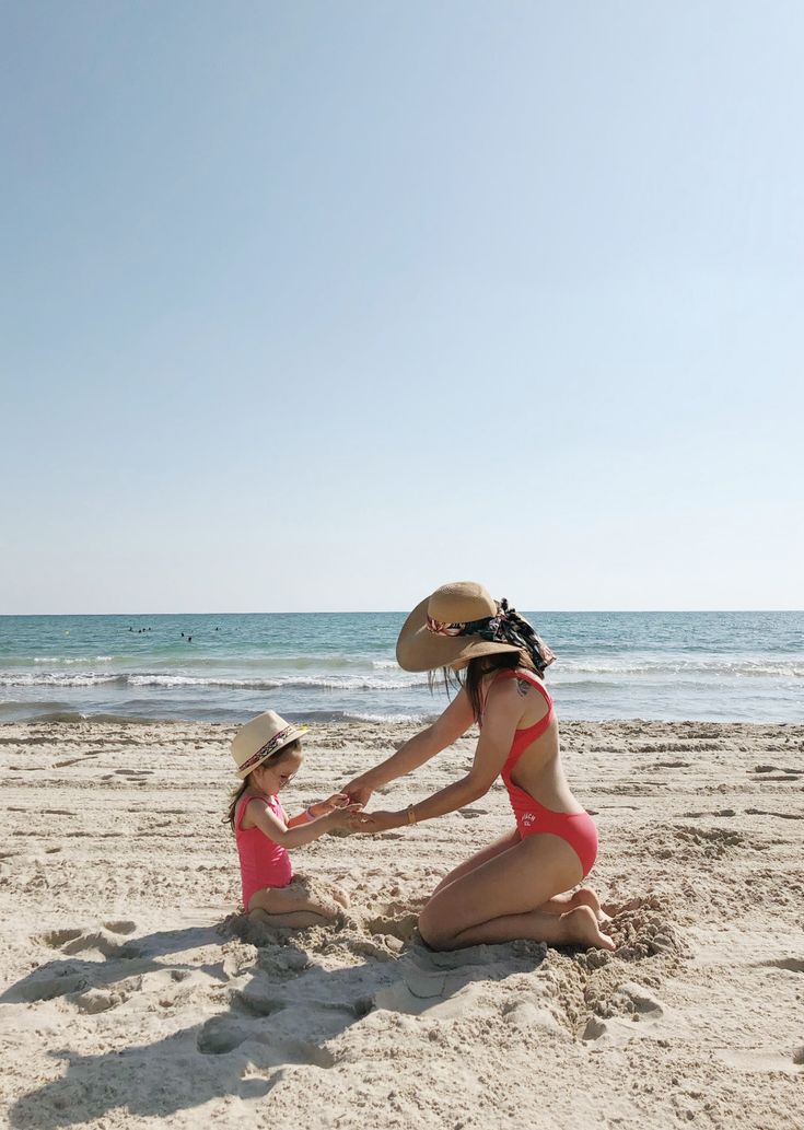 two women in bikinis and hats sitting on the sand at the beach, touching hands with each other