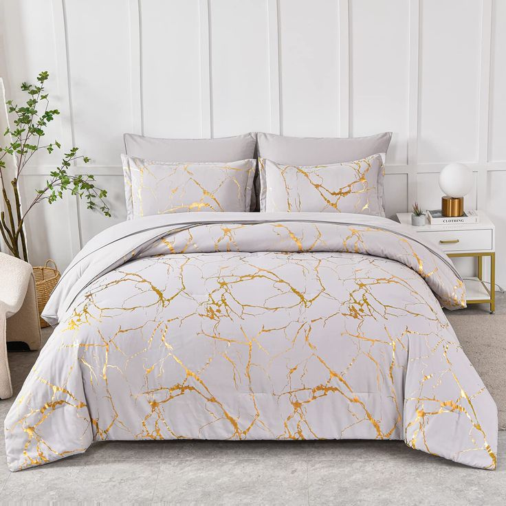 a bed with white and gold comforters in a room next to a plant on the floor