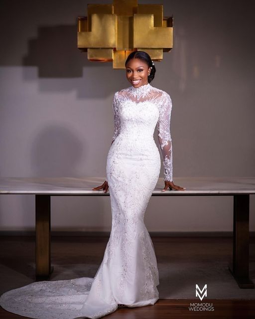 a woman standing in front of a table wearing a white wedding dress with long sleeves