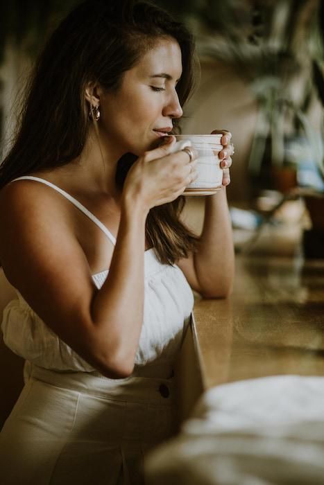 a woman in a white dress drinking out of a coffee cup with her eyes closed