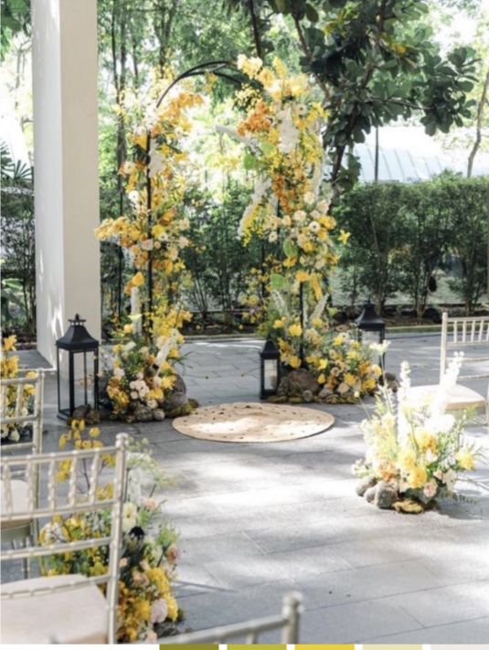 an outdoor wedding venue with white chairs and yellow flowers on the arch, surrounded by greenery