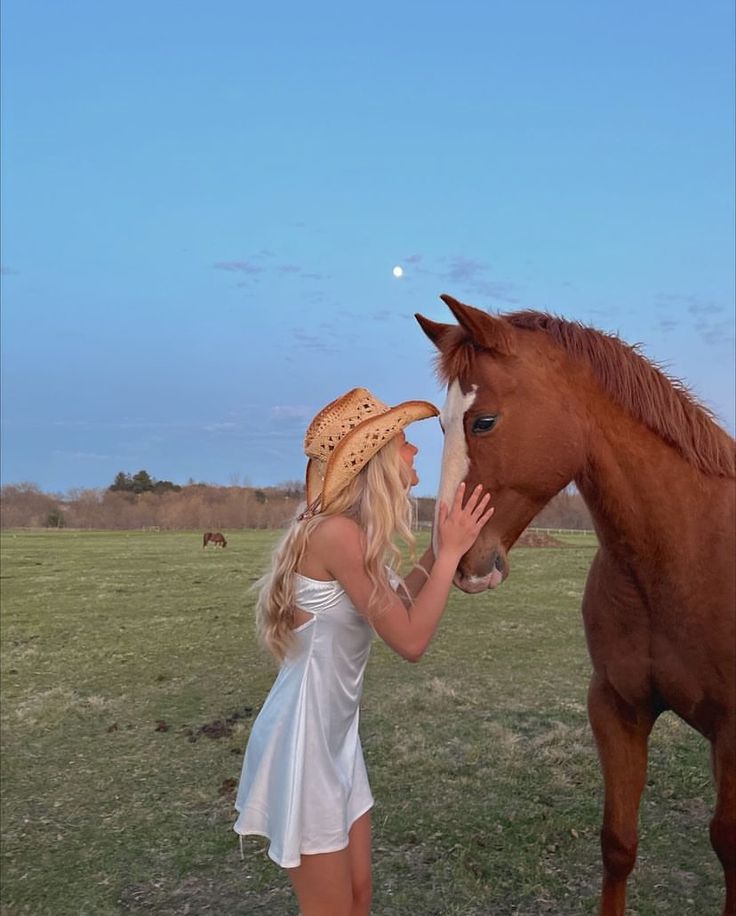 a woman standing next to a brown horse on top of a lush green field under a blue sky