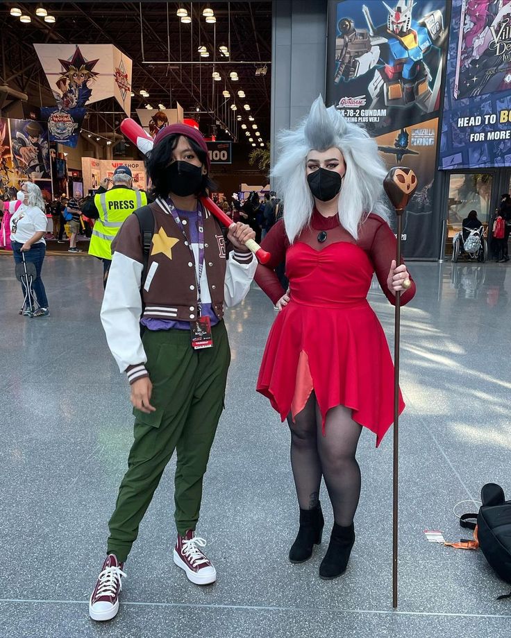 two people in costumes standing next to each other at an indoor convention center, one wearing a mask and the other holding a stick