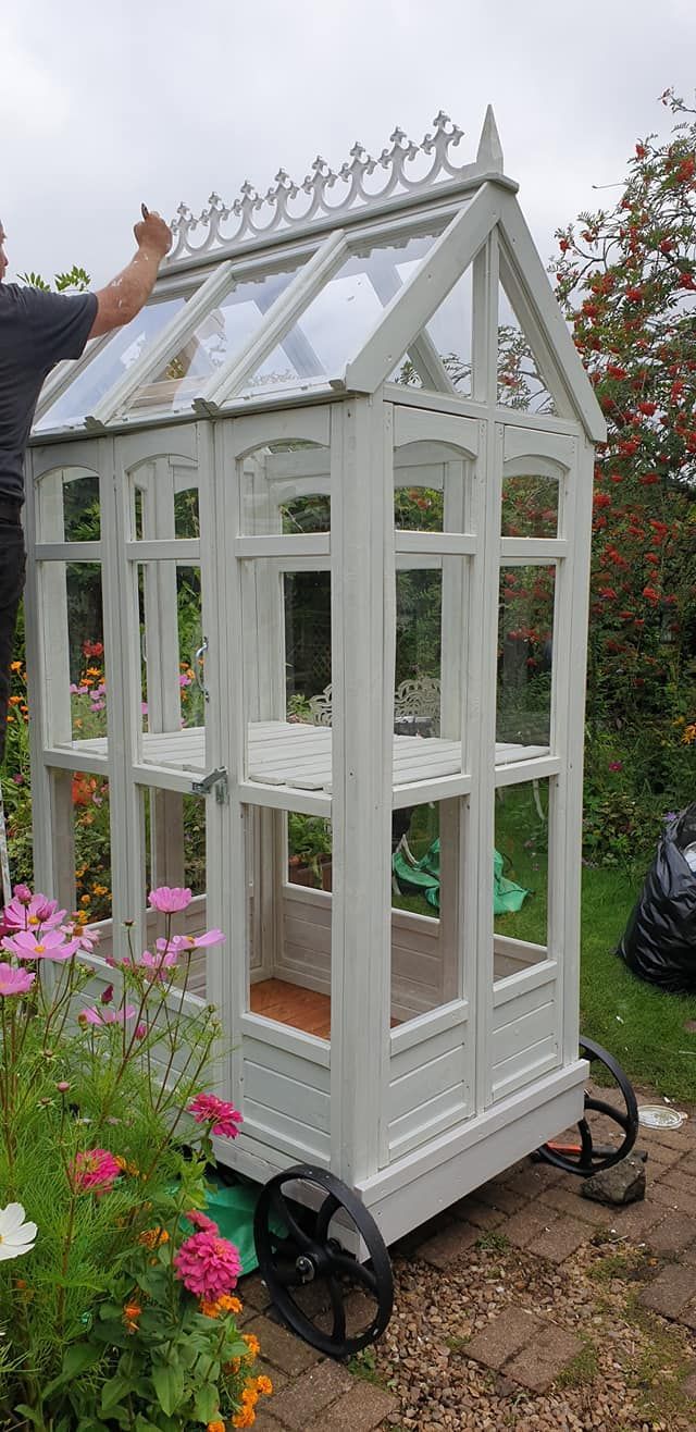 a man standing next to a small white greenhouse with flowers in the front and behind it