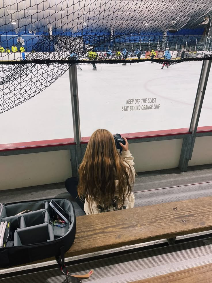 a woman sitting on a bench taking a photo with her cell phone in front of an ice rink