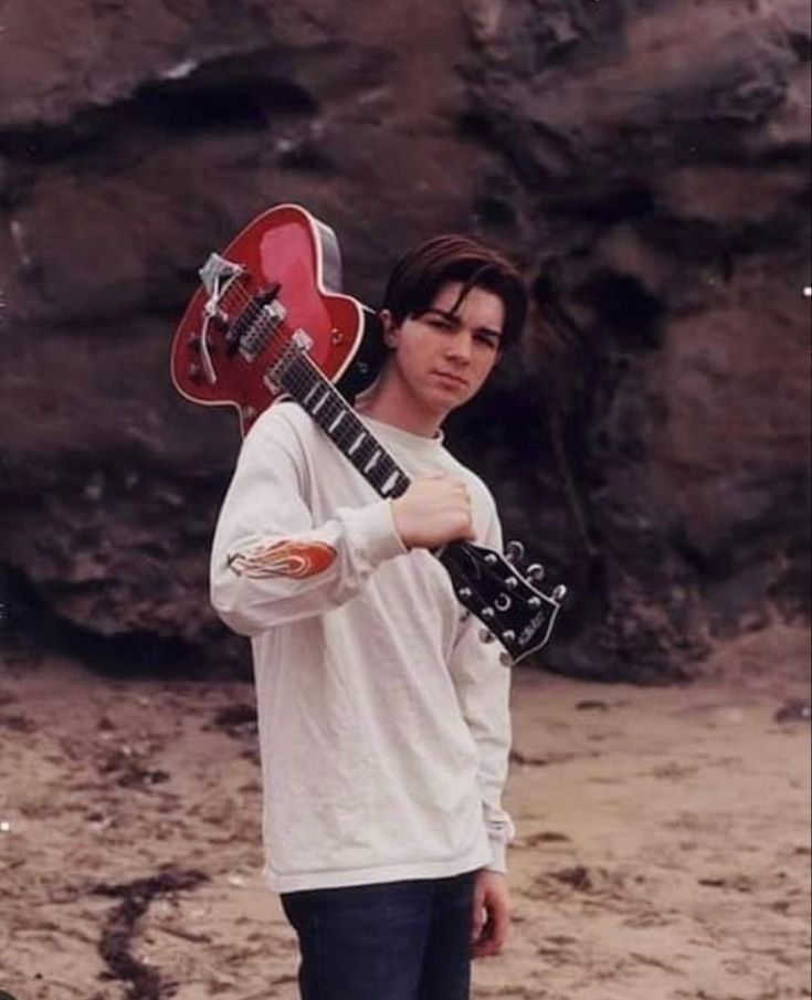 a young man holding a red guitar in his right hand while standing on the beach