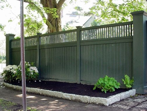 a green fence with white flowers and trees in the back ground, next to a brick walkway