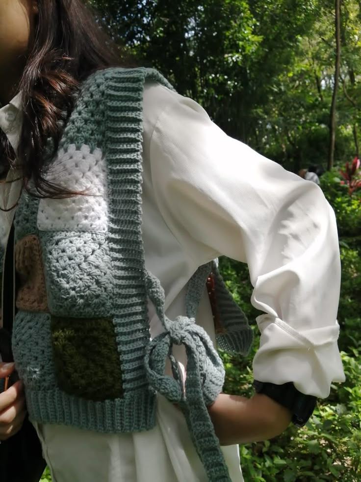 a woman wearing a green crocheted vest and white dress in front of trees