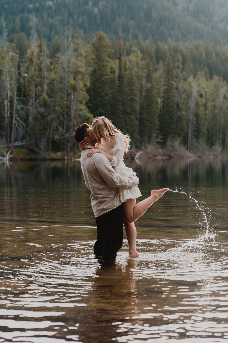 a man and woman are kissing in the water near a lake surrounded by pine trees