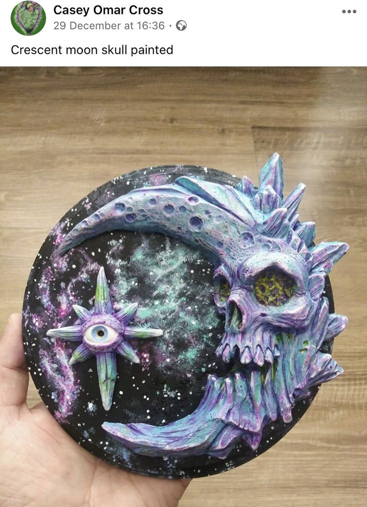 a hand holding up a purple and blue moon with two stars on the bottom, and a skull in the middle