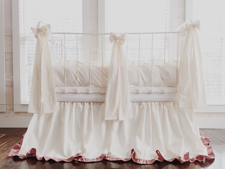 a white crib with pink ruffles and bows on the bed skirt, in front of large windows