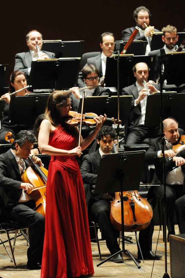 a woman in a red dress is playing the violin with orchestra members behind her on stage