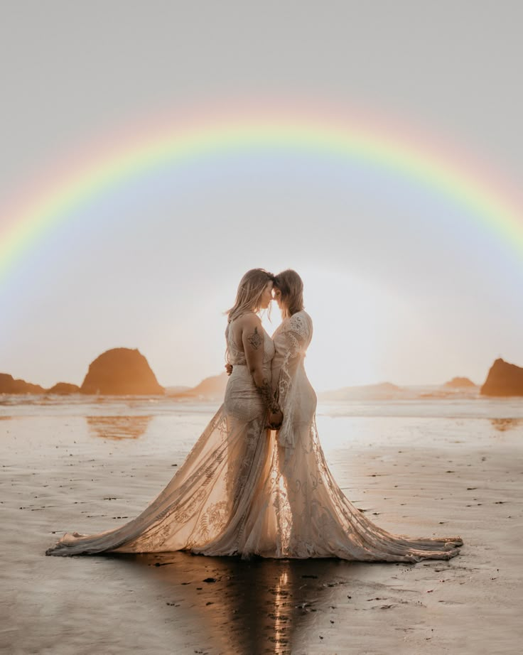 two women in white dresses standing on the beach under a rainbow