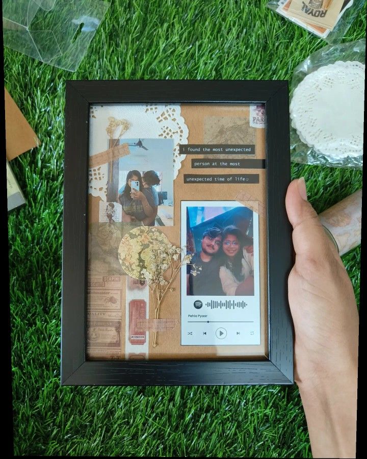 a person holding up a framed photo in the grass with flowers and other items around it