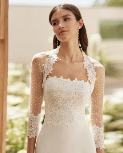 a woman wearing a white dress with sheer sleeves and pearls on the neck, standing in front of flowers