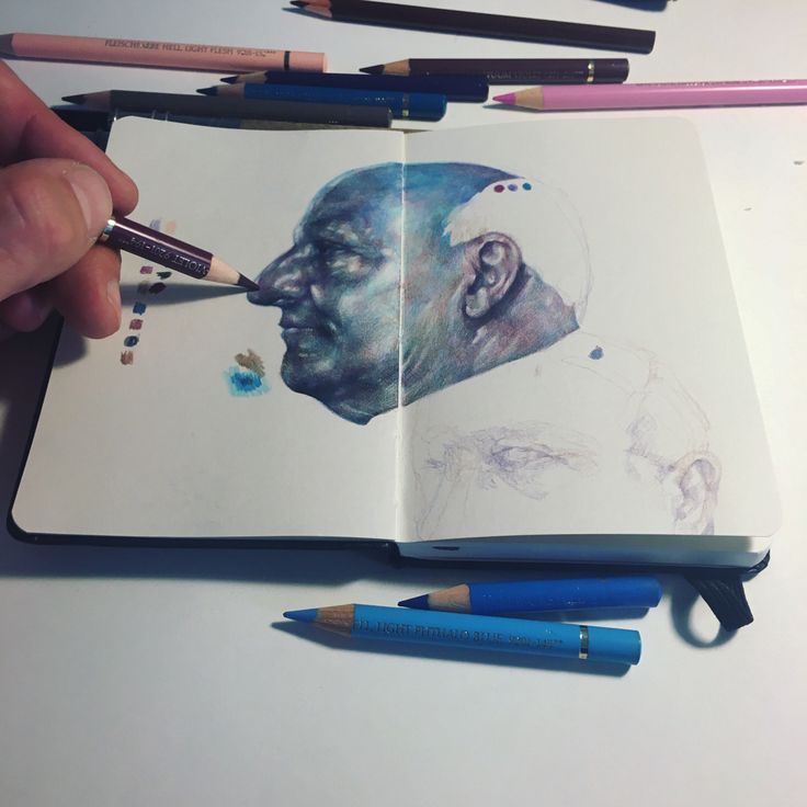 a hand is holding a pencil over an open book with drawings on it and two colored pencils next to it