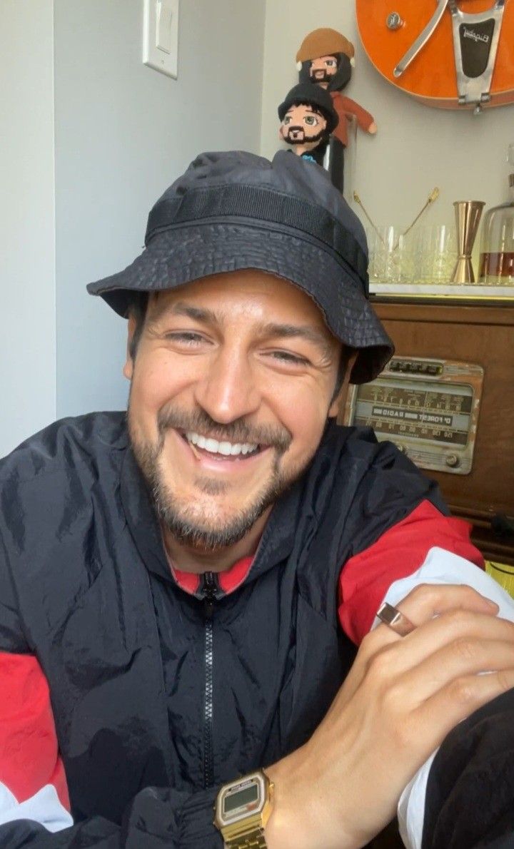 a man wearing a hat and jacket sitting in front of a wall with clocks on it