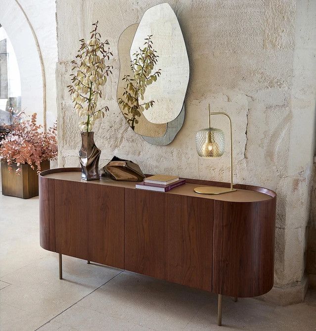 a wooden sideboard sitting next to a mirror and vase with flowers on it in front of a stone wall