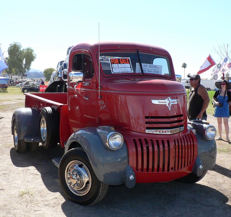 an old red truck parked on top of a dirt field next to other cars and people