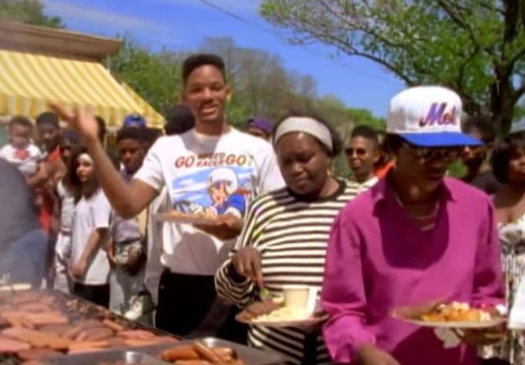 a group of people standing around a bbq with hot dogs on top of it