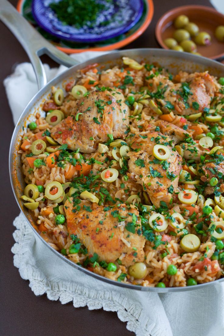 a pan filled with chicken and vegetables on top of a table next to bowls of olives