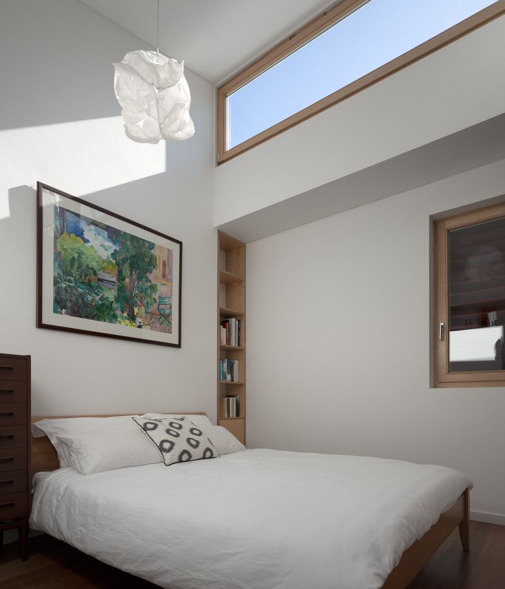 a white bed sitting under a window next to a wooden shelf filled with books on top of a hard wood floor