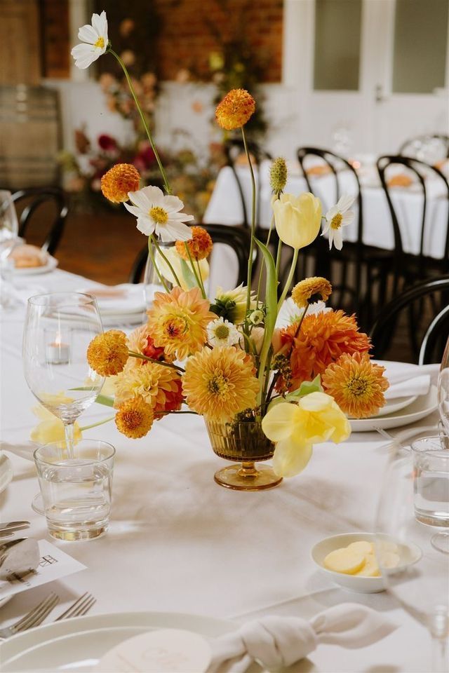 an arrangement of flowers in a vase on a table with white linens and place settings