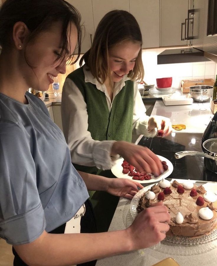 two women in the kitchen decorating a cake with icing and strawberries on it