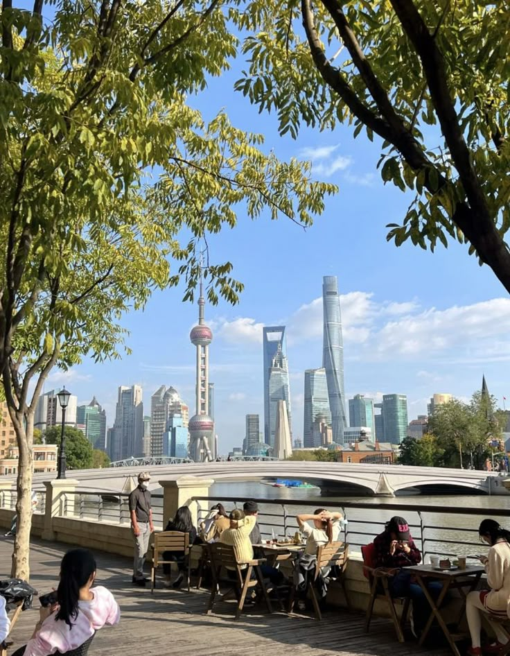 people are sitting on benches near the water in front of some tall buildings and trees