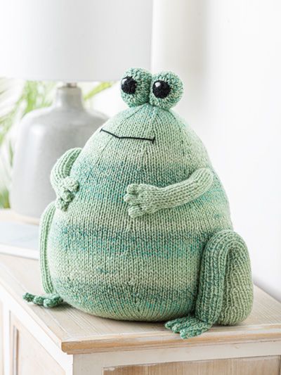 a green stuffed frog sitting on top of a wooden table next to a white lamp