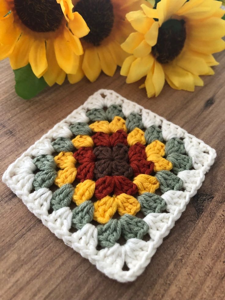 a crocheted square sitting on top of a wooden table next to sunflowers