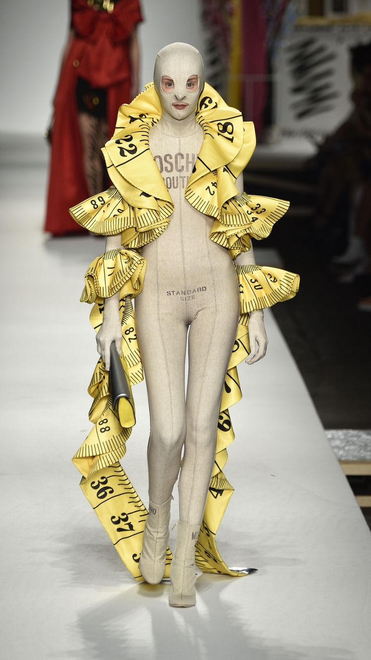 a white mannequin with yellow ribbons and numbers on it's body, walking down a runway