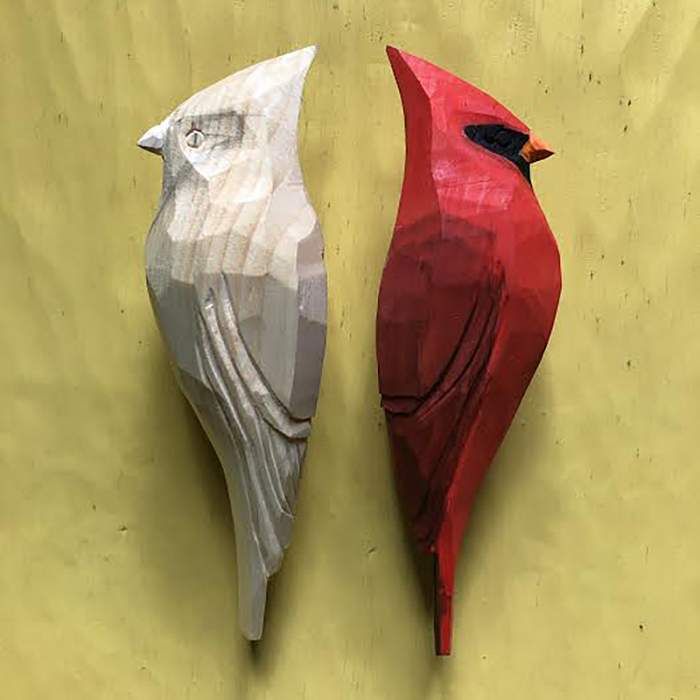 two red and white birds sitting next to each other on a yellow surface, one is made out of wood