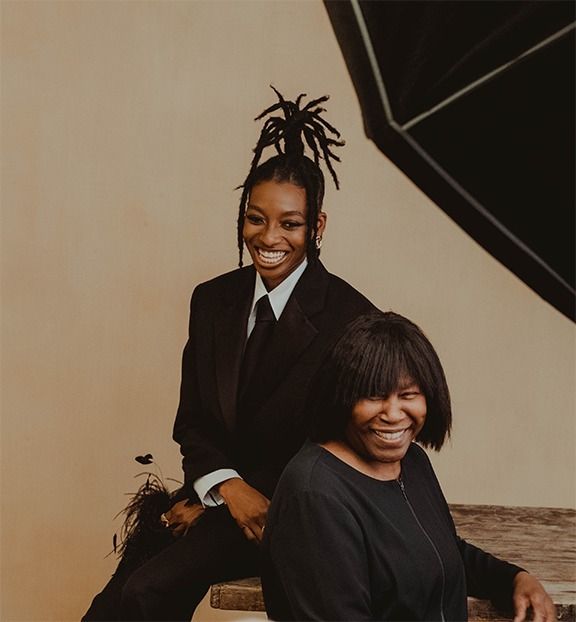 two women are sitting on a bench and one is wearing a black suit with dreadlocks