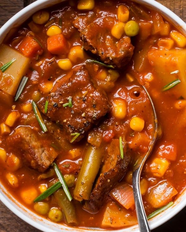 a bowl filled with meat and vegetables on top of a wooden table next to a spoon