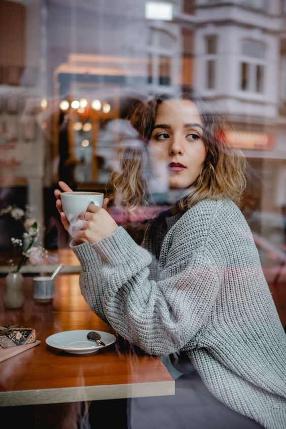 a woman sitting at a table with a cup in her hand looking out the window