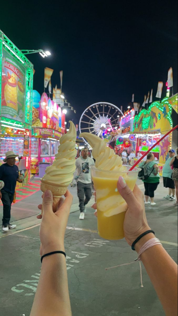 two people holding up ice cream cones in front of carnival rides and ferris wheel at night