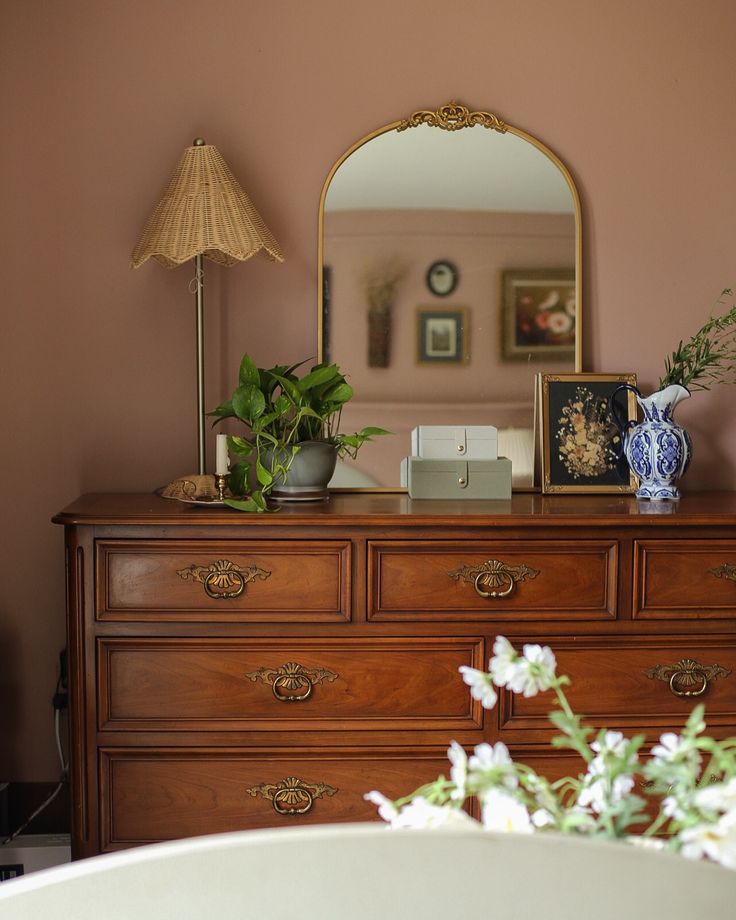 a dresser with flowers and pictures on it in front of a mirror, lamp and vase