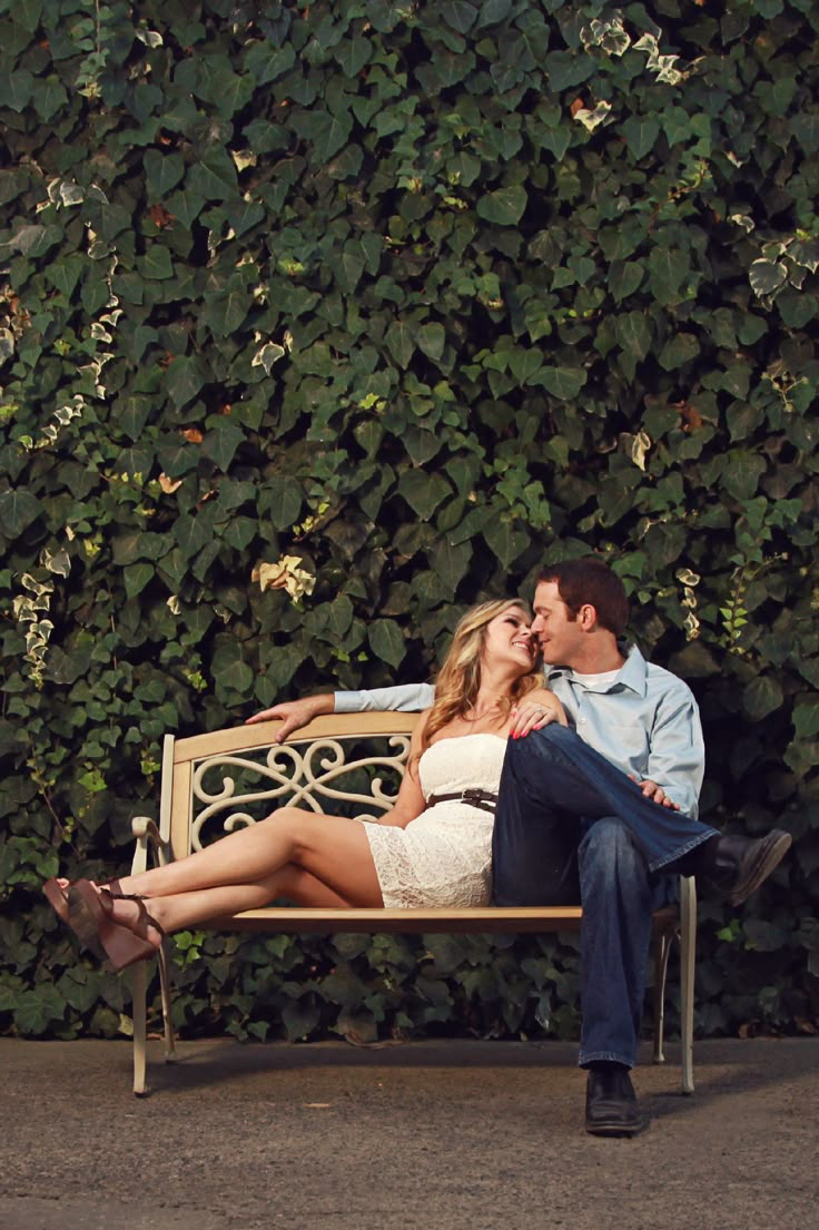 a man and woman sitting on a bench in front of a wall with green plants