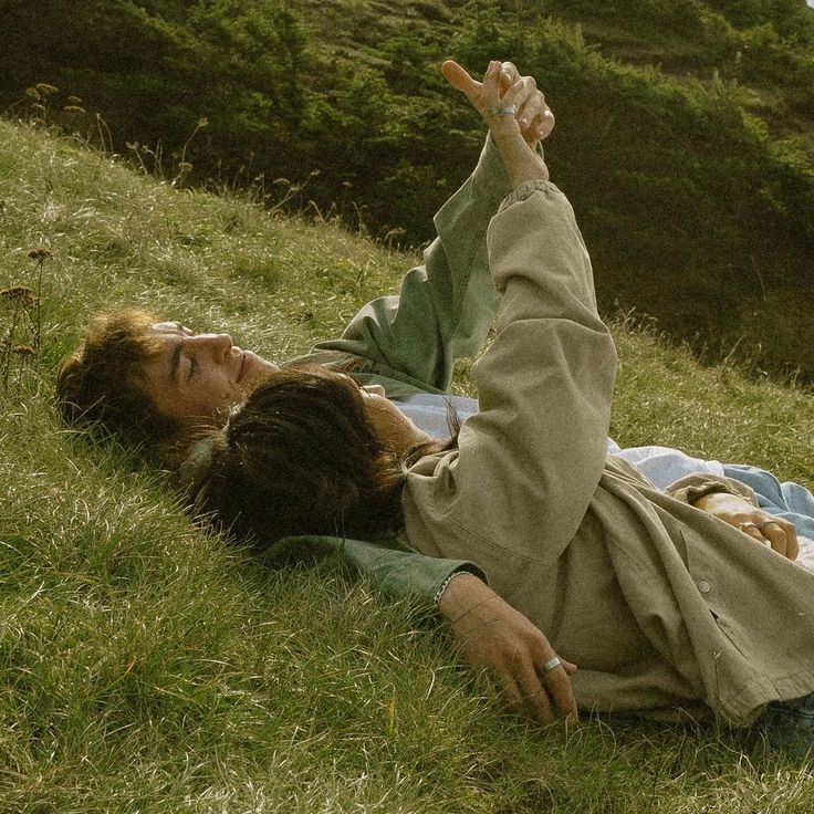 two people laying in the grass on top of a hill with one pointing at something