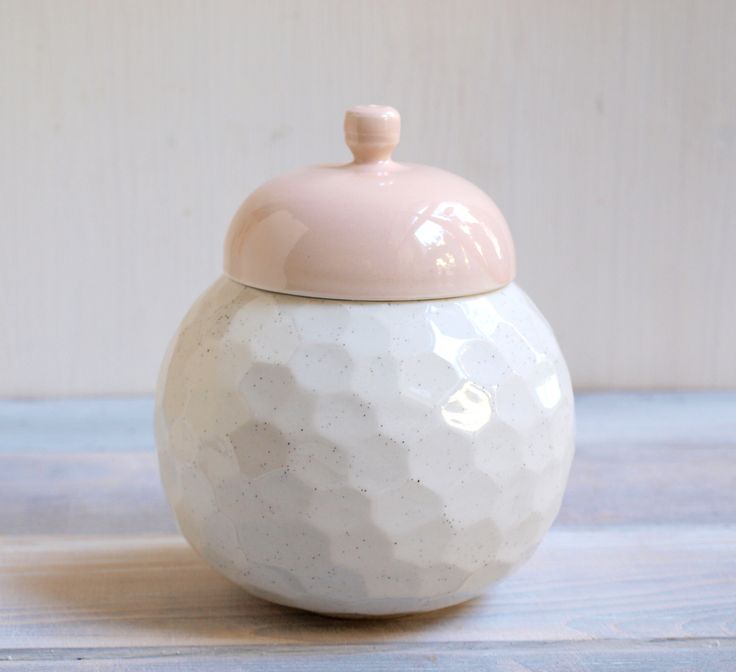 a white and pink ceramic jar with a lid on a wooden table next to a wall