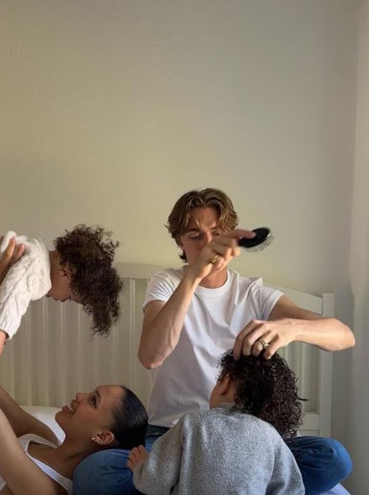 four people sitting on top of a bed with one person brushing their hair and the other holding a remote control