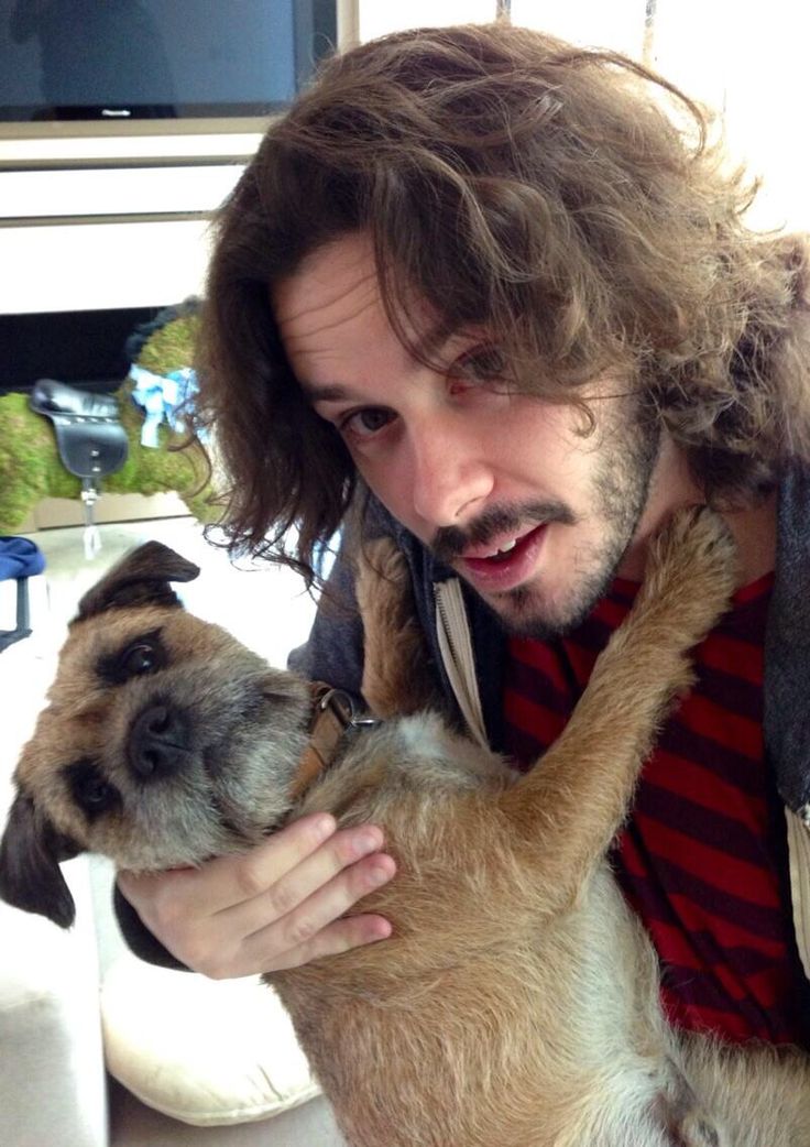 a man with long hair holding a dog in his arms and looking at the camera