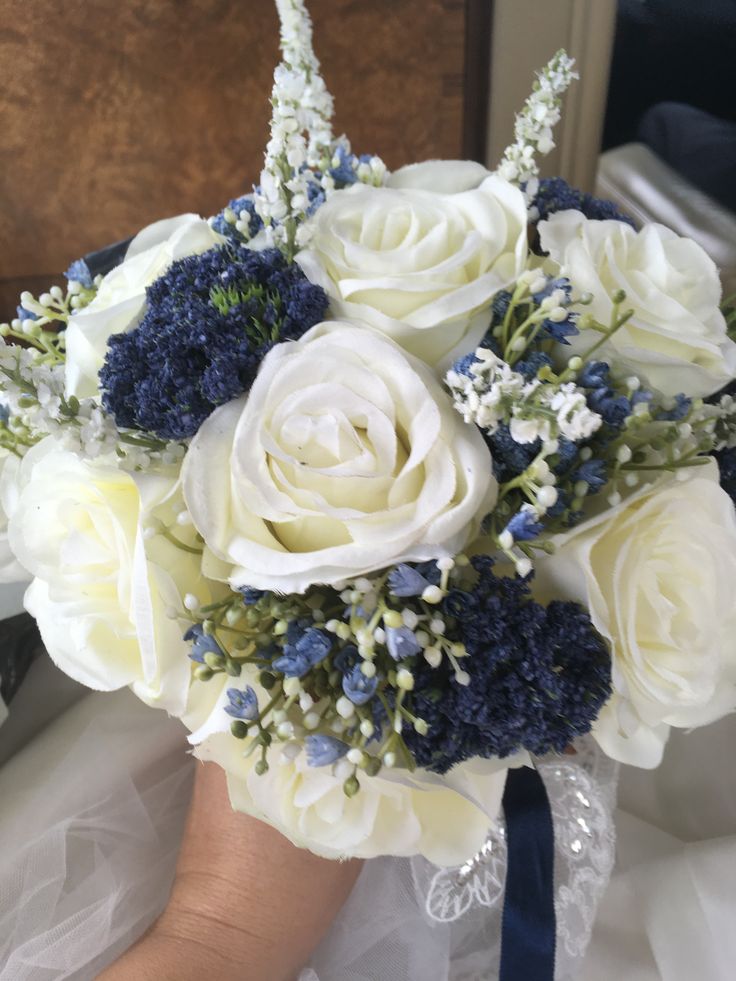 a bridal bouquet with white roses and blue hydrangeas is held by a bride's hand