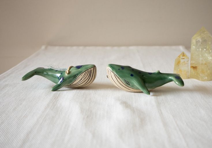 two ceramic dolphins sitting next to each other on a white tablecloth with a yellow rock in the background