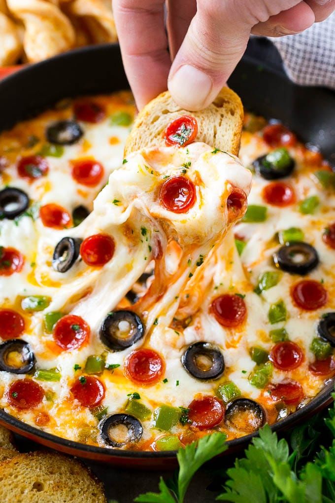 a hand dipping a piece of pizza into a skillet with olives and pepperoni