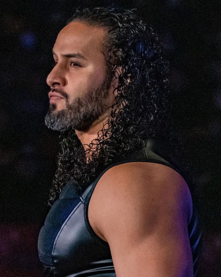 a man with long hair and beard wearing a black wrestling outfit standing in front of a crowd