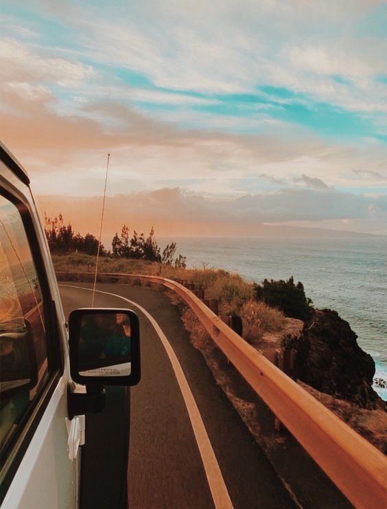 a car driving down a road next to the ocean at sunset or sunrise with clouds in the sky